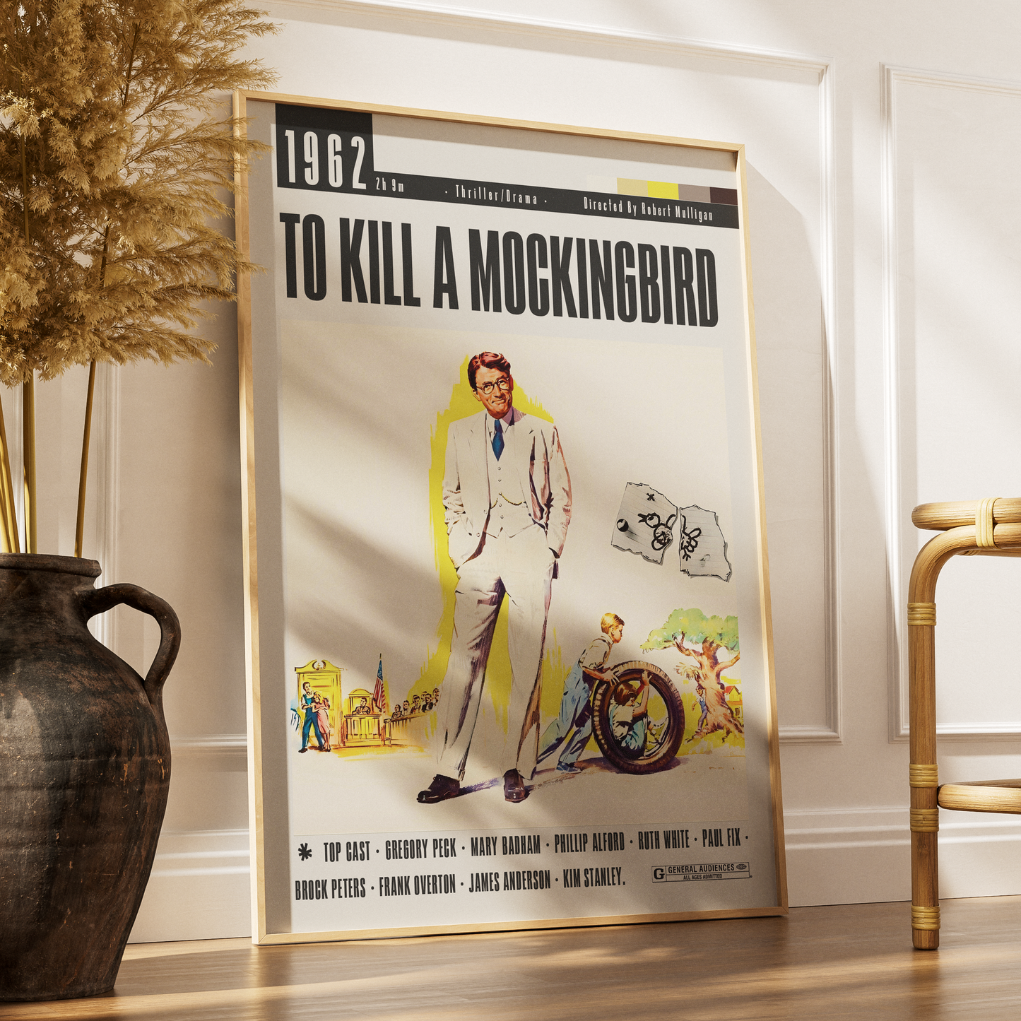 A framed vintage movie poster for 'To Kill a Mockingbird' leaning against a white wall in a styled interior setting with a dark vase and dried grass on the left and a bamboo chair on the right.
