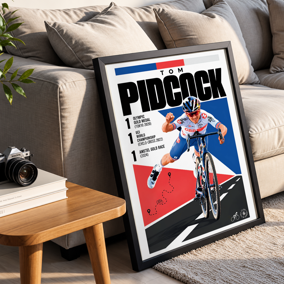 A framed cycling poster featuring Tom Pidcock leaning against a beige sofa in a living room setting, with a small wooden side table and camera in the foreground.