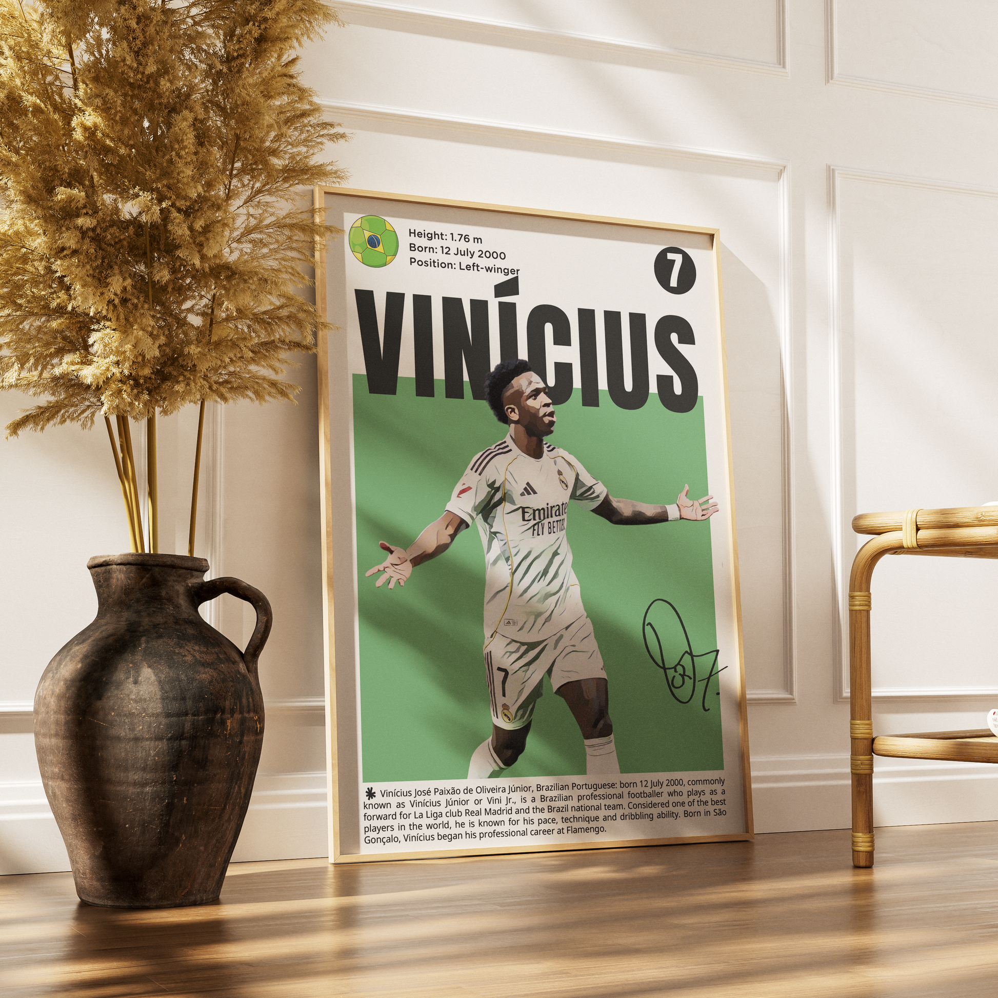 A framed football poster of Vinícius Jr. leaning against a white paneled wall on a wooden floor, styled with a dark ceramic vase containing dried pampas grass on the left and a gold-framed chair on the right.