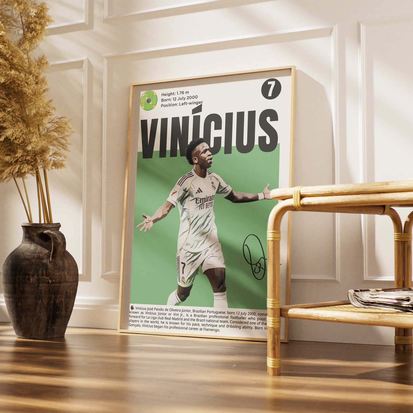 A framed soccer poster of Vinicius Jr. leaning against a white paneled wall on a wooden floor, next to a bamboo side table and a dark ceramic vase with dried grass.