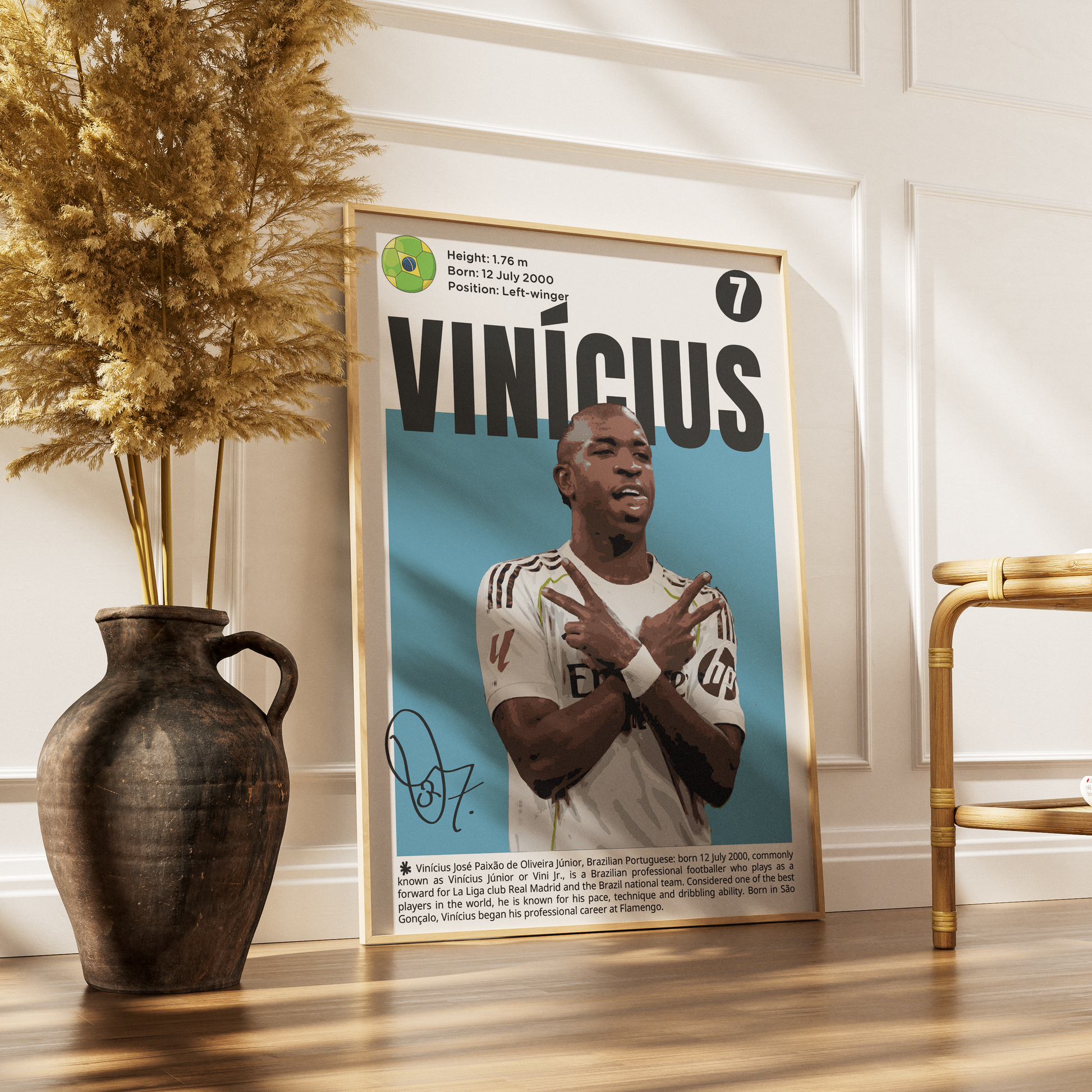 A framed poster featuring a portrait of soccer player Vinícius with the name 'VINÍCIUS' in large letters, styled leaning against a white paneled wall on a wooden floor beside a dark vase with dried grass and a gold chair.