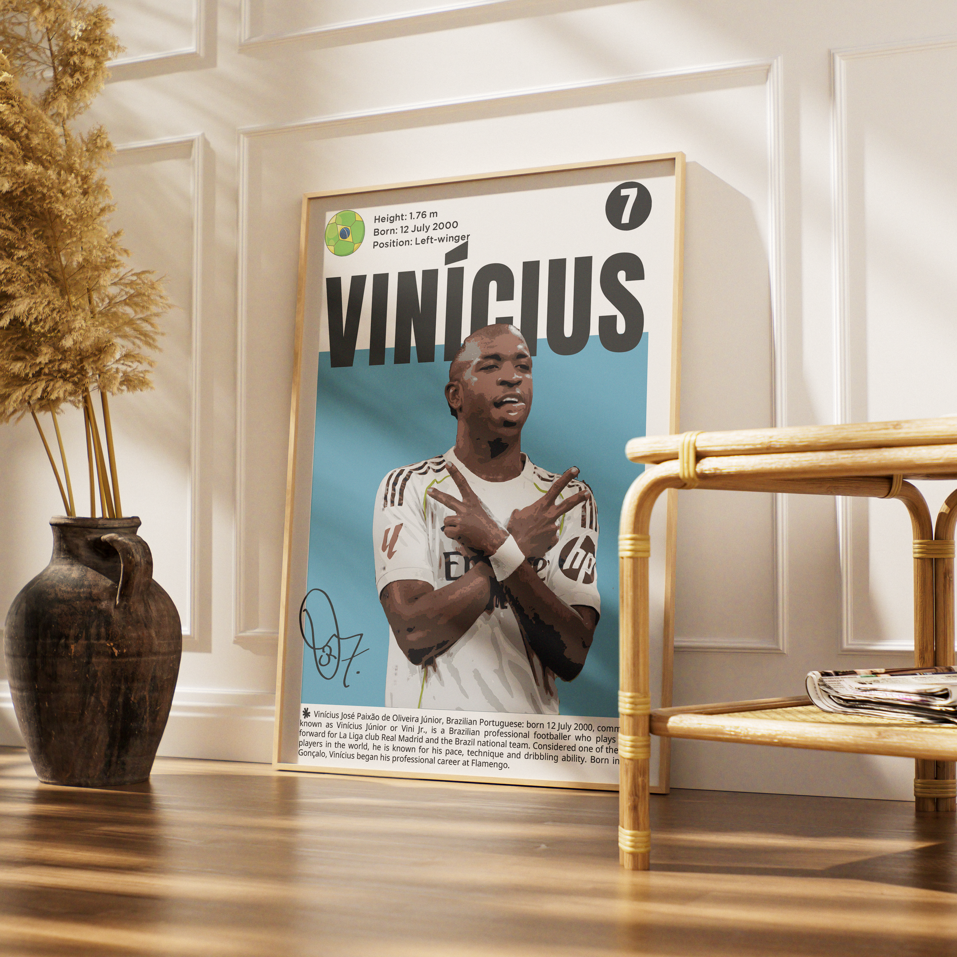 A framed poster of soccer player Vinicius Jr. leaning against a white paneled wall in a styled room with a wooden floor, a dark vase with dried grass, and a bamboo side table.