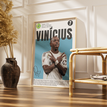 A framed poster of soccer player Vinicius Jr. leaning against a white paneled wall in a styled room with a wooden floor, a dark vase with dried grass, and a bamboo side table.