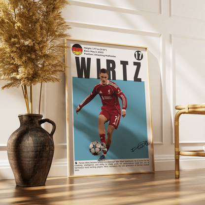 A framed poster featuring a football player in a red Liverpool jersey with the name 'WIRTZ' and biographical text, leaning against a white paneled wall on a wooden floor next to a dark vase with dried grass and a gold chair.