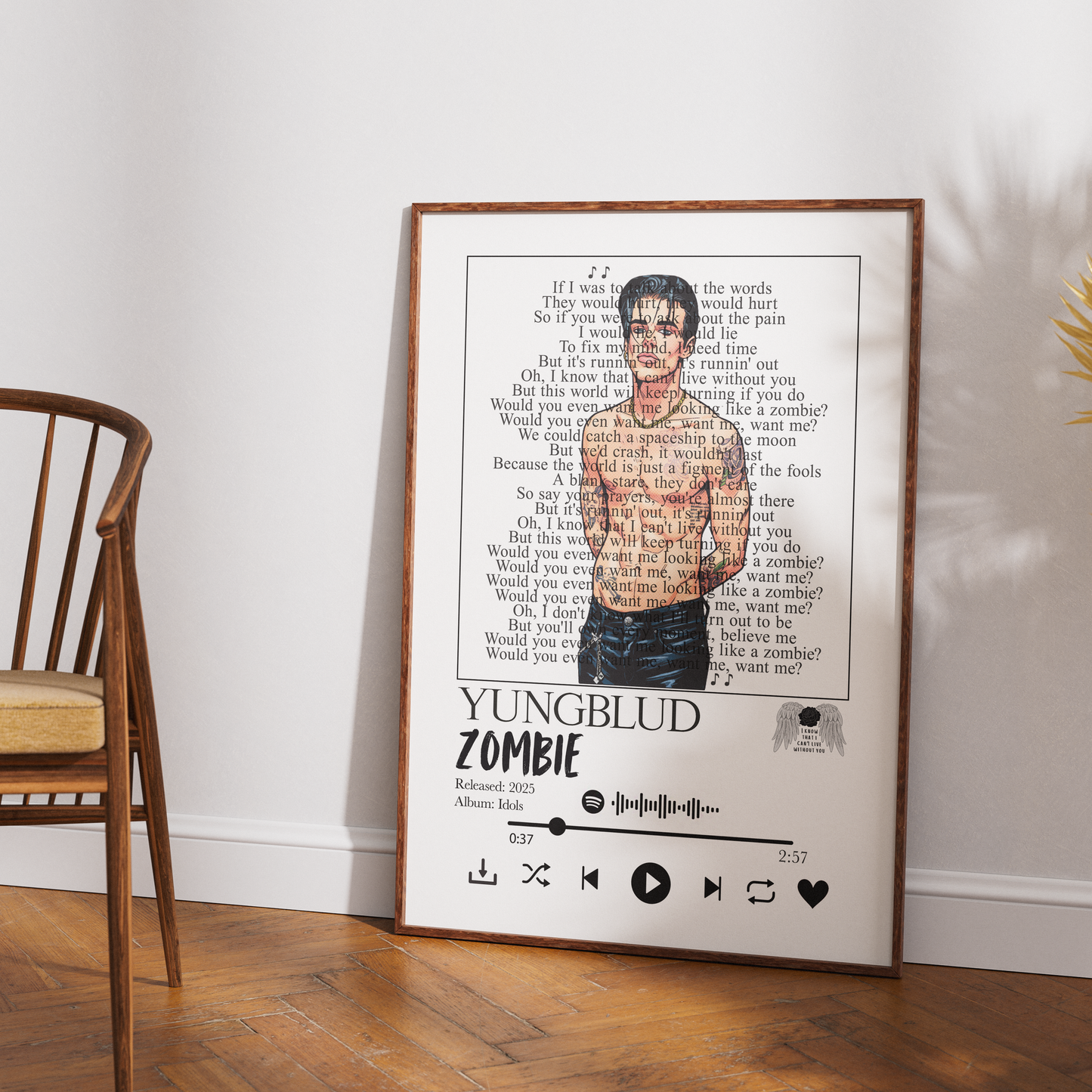 A framed poster print leaning against a white wall on a wooden floor, featuring song lyrics with a portrait of Yungblud and the text 'YUNGBLUD ZOMBIE' and 'Referenced 2005; Album: I'm Good at Losing You.' A wooden chair is visible on the left, and a plant leaf appears on the right.