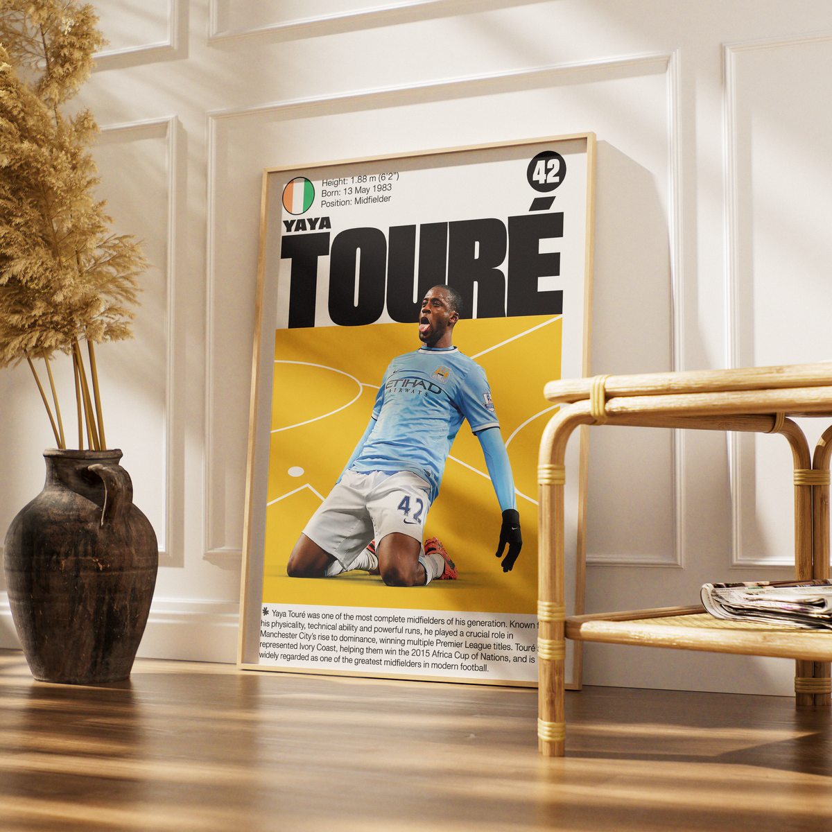 A framed sports poster of Yaya Touré from his 2005 stint with Manchester City, styled in a warm, modern interior setting next to a bamboo side table and a dark ceramic vase with dried grass.