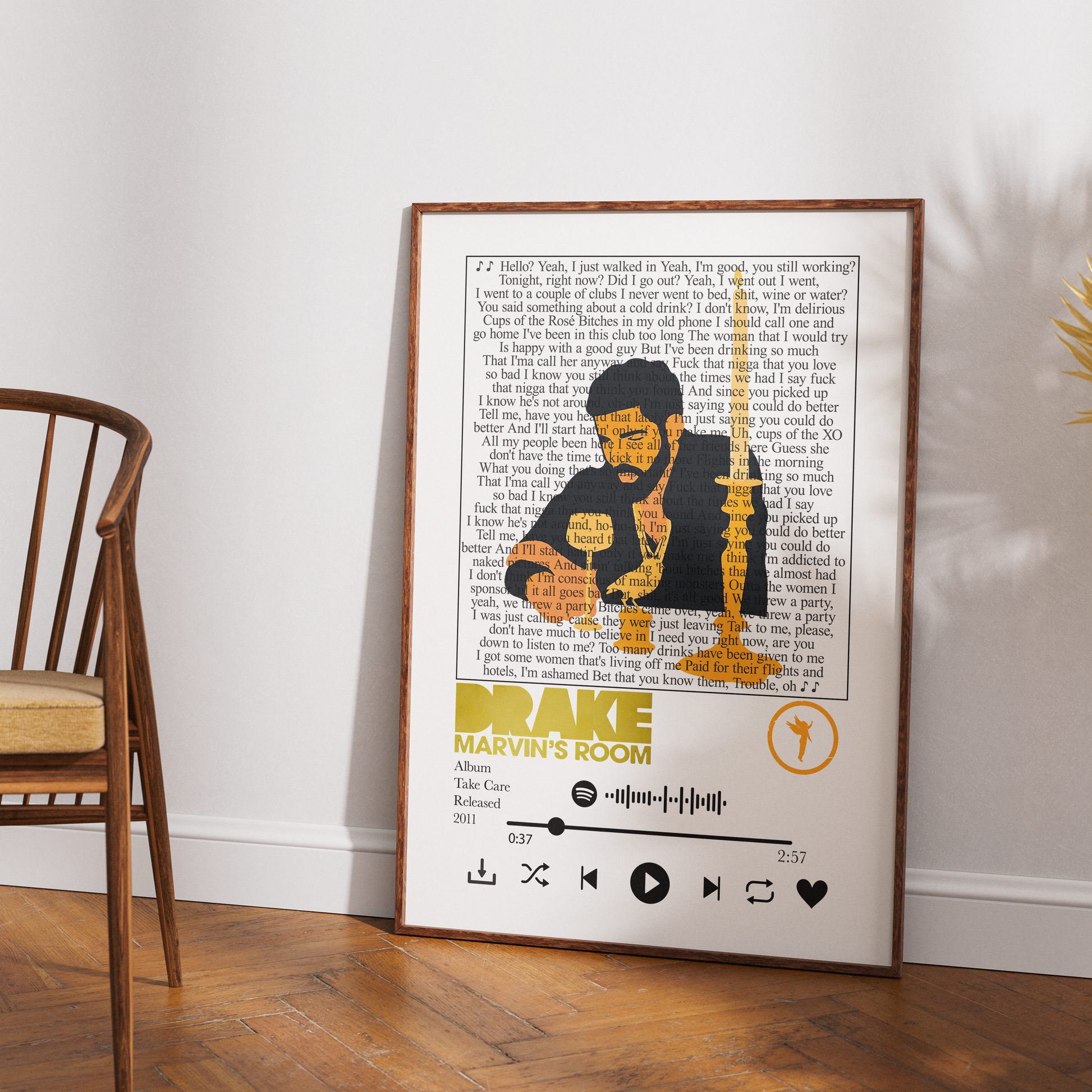 A framed art print featuring a stylized portrait of Drake with song lyrics and the text 'Drake - Marvin's Room' in a music player design, leaning against a white wall on a wooden floor next to a chair.