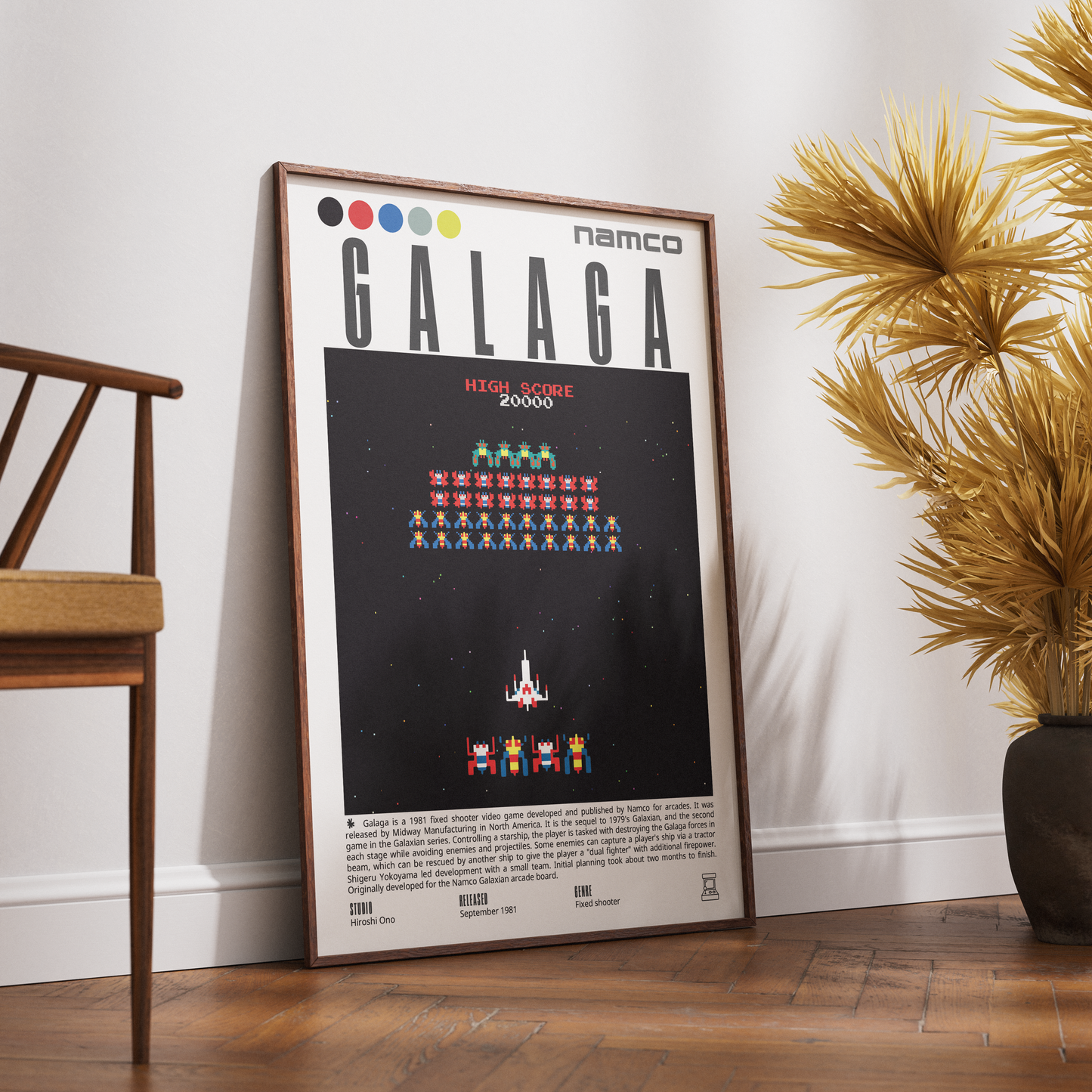 A framed vintage-style poster leaning against a white wall on a wooden floor, next to a chair and a potted palm. The poster features the Namco GALAGA game title with pixel-art characters on a black background.