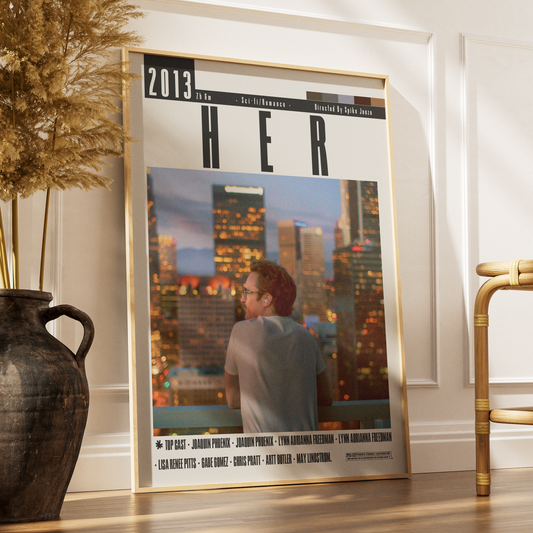A framed poster leaning against a white wall on a wooden floor, next to a dark vase with dried grass and part of a gold chair. The poster features a magazine-style cover with the title 'HER' and a photo of a person in front of a city skyline at dusk.