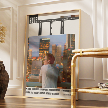 A large framed poster leaning against a white paneled wall in a styled interior. The poster features the title 'HER' with '2013' at the top and a photo of a man looking out over a city skyline at dusk.