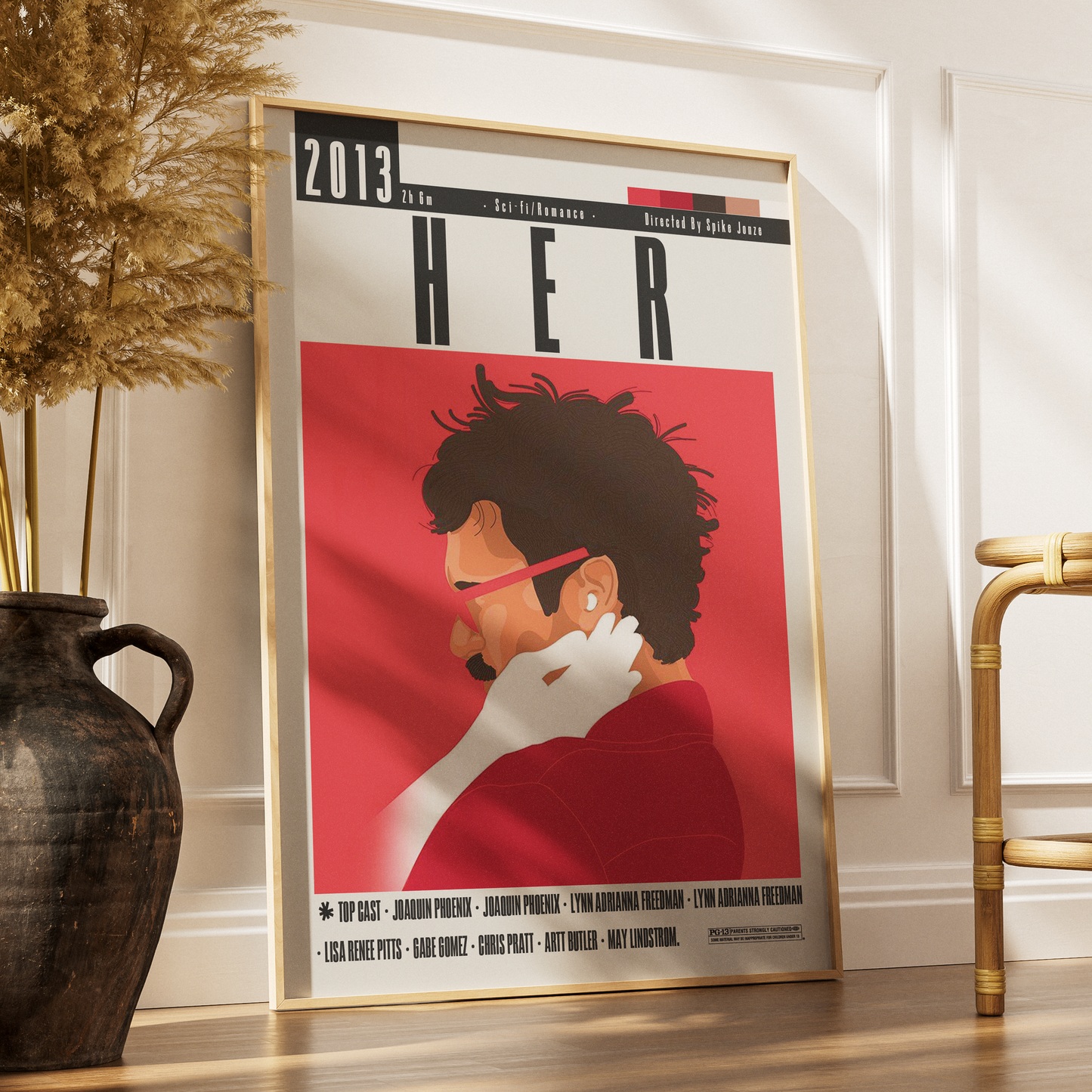 A framed movie poster for the 2013 film 'Her' leaning against a white wall on a wooden floor, styled in a modern room with a dark ceramic vase and dried grass on the left and a gold chair on the right.