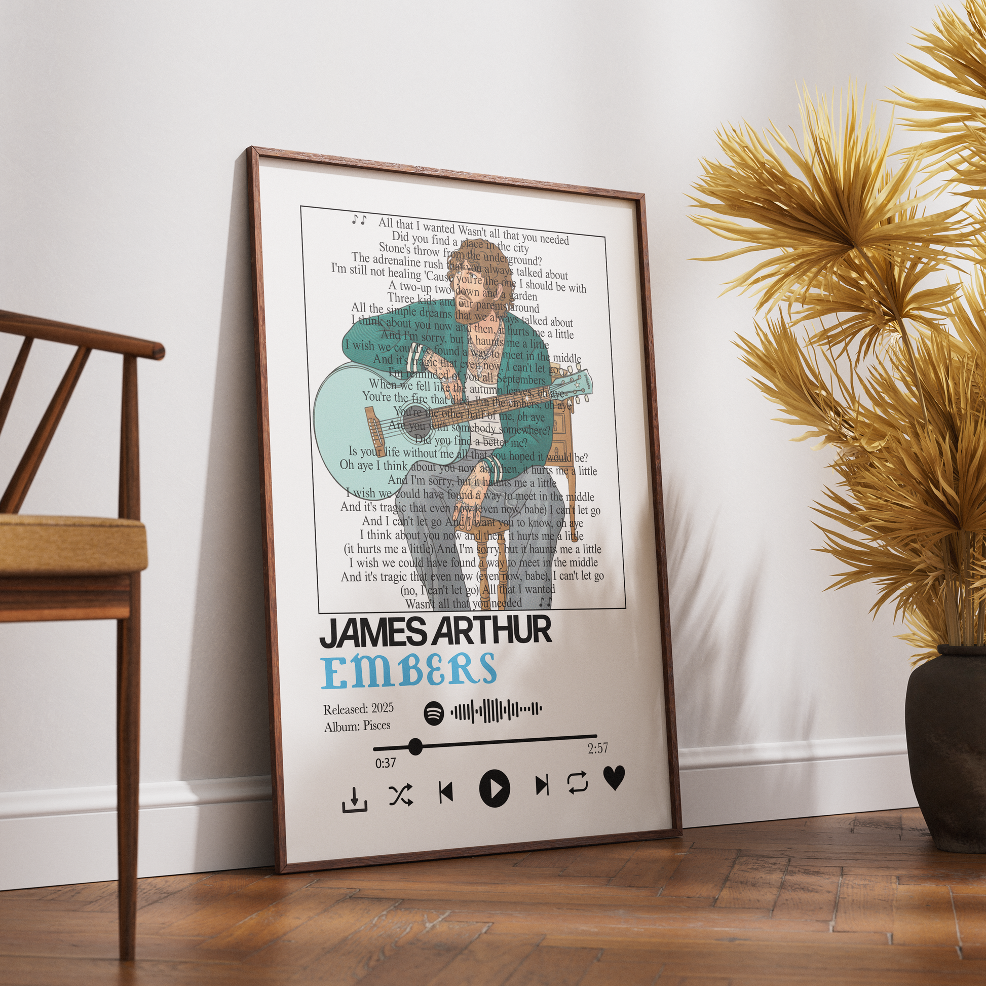 A framed music-themed poster of James Arthur Embers leaning against a white wall on a wooden floor, styled in a room next to a wooden chair and a potted plant.
