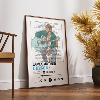 A framed music-themed poster of James Arthur Embers leaning against a white wall on a wooden floor, styled in a room next to a wooden chair and a potted plant.