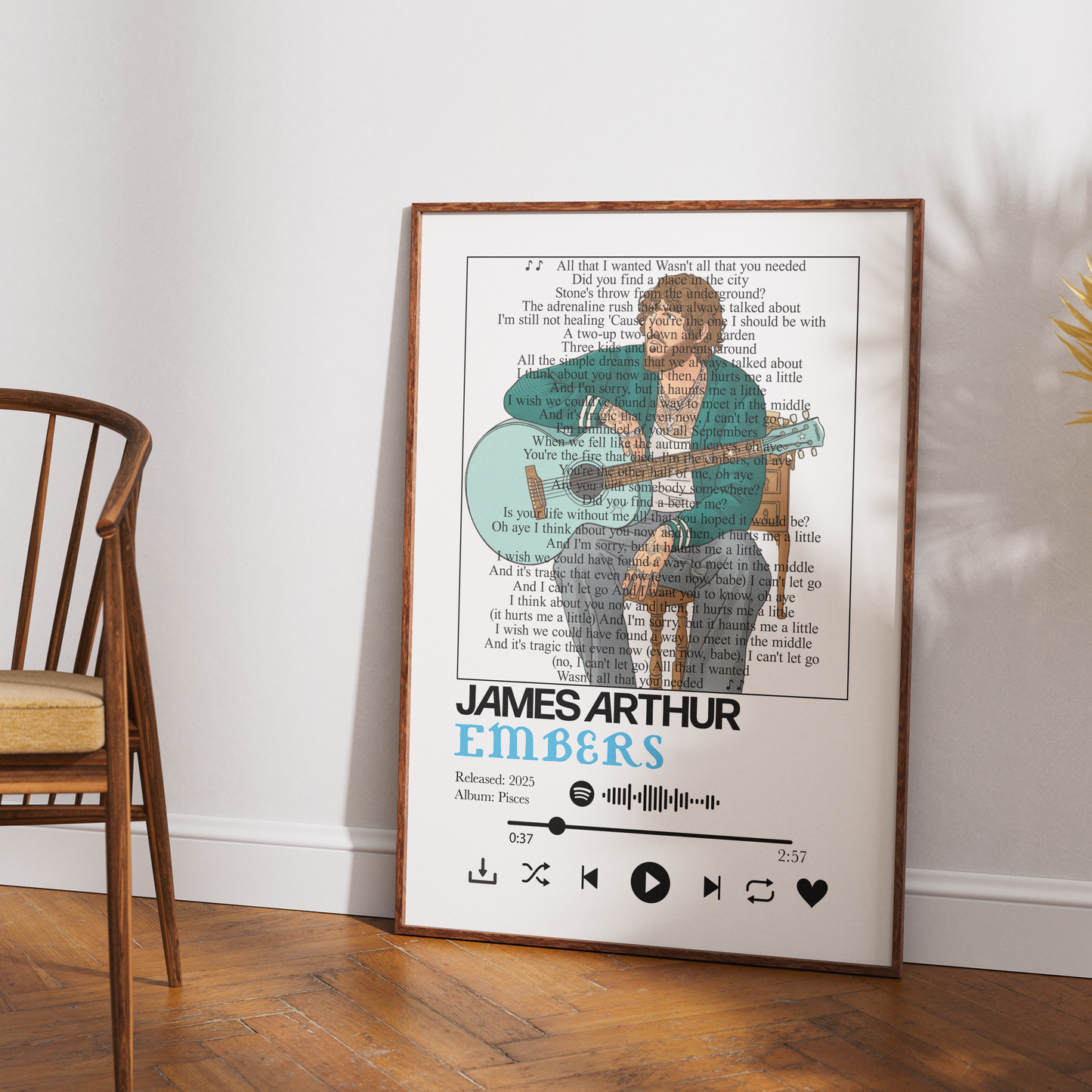 A framed personalized song poster featuring James Arthur Embers, styled leaning against a white wall on a wooden floor next to a wooden chair.