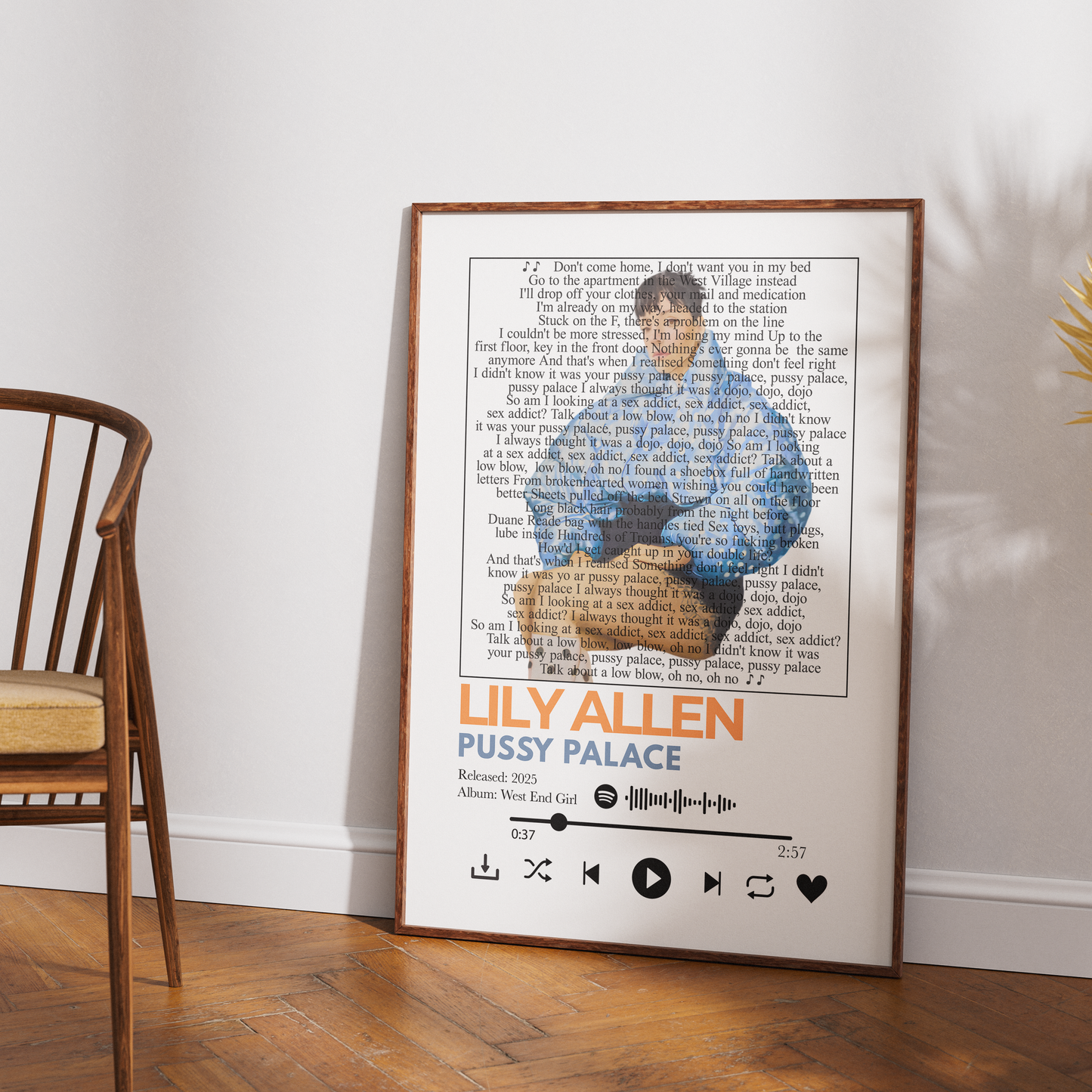 A framed music poster of Lily Allen's album 'Pussy Palace' leaning against a white wall on a wooden floor, next to a wooden chair.