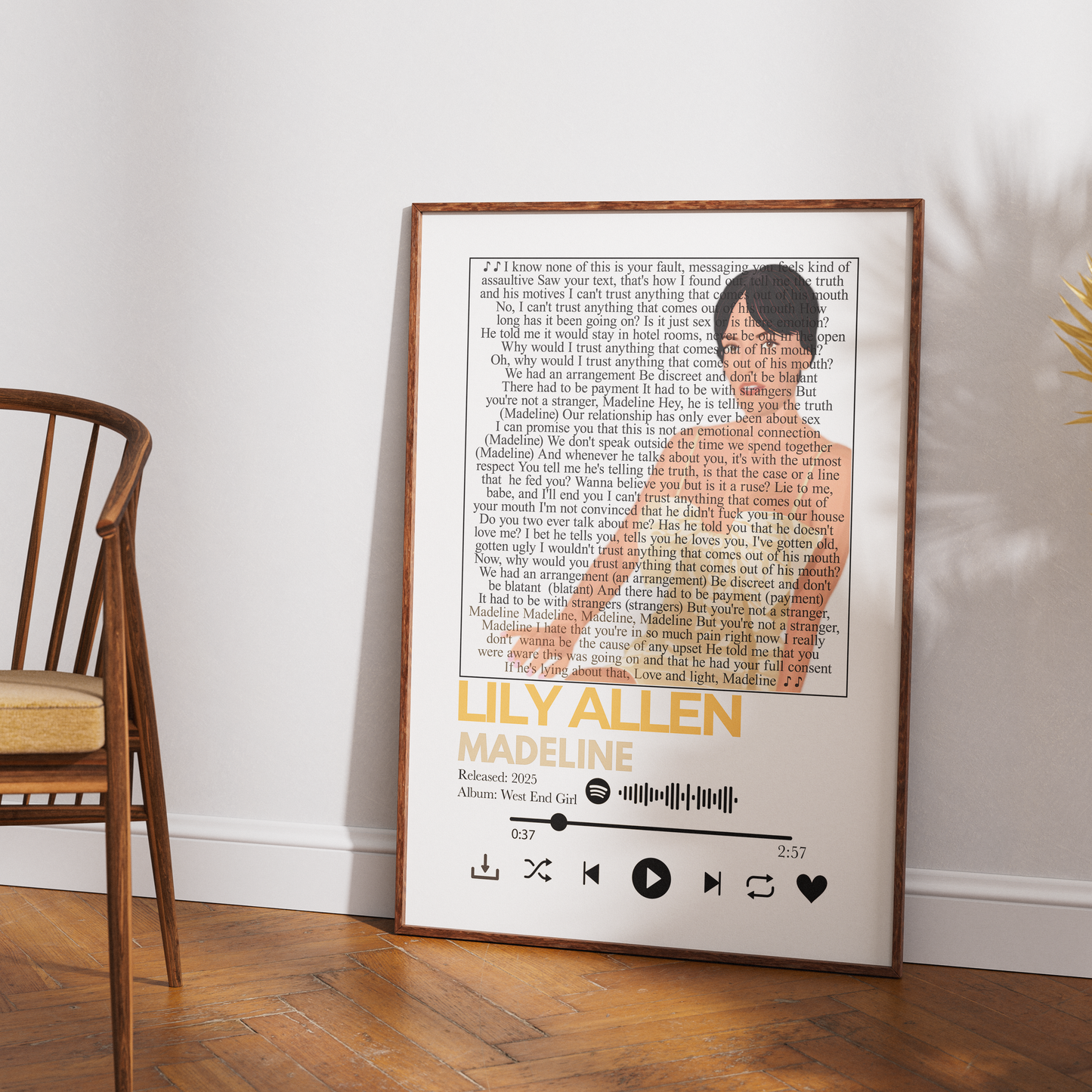 A framed music-themed art print leaning against a white wall on a wooden floor, next to a wooden chair. The print features a song lyric background with a silhouette of Ulysses S.