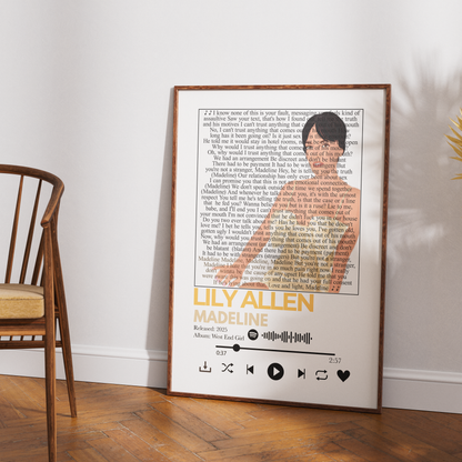 A framed music-themed art print leaning against a white wall on a wooden floor, next to a wooden chair. The print features a song lyric background with a silhouette of Ulysses S.