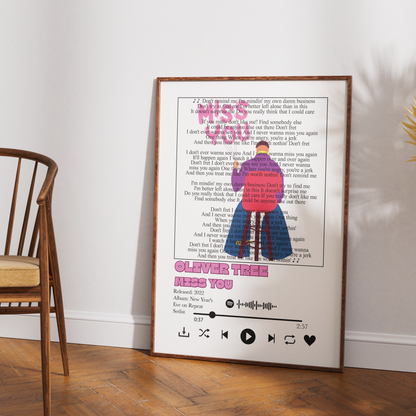 A framed custom song print leaning against a white wall on a wooden floor, next to a wooden chair. The print features a book page background with the text 'Oliver Tree N.