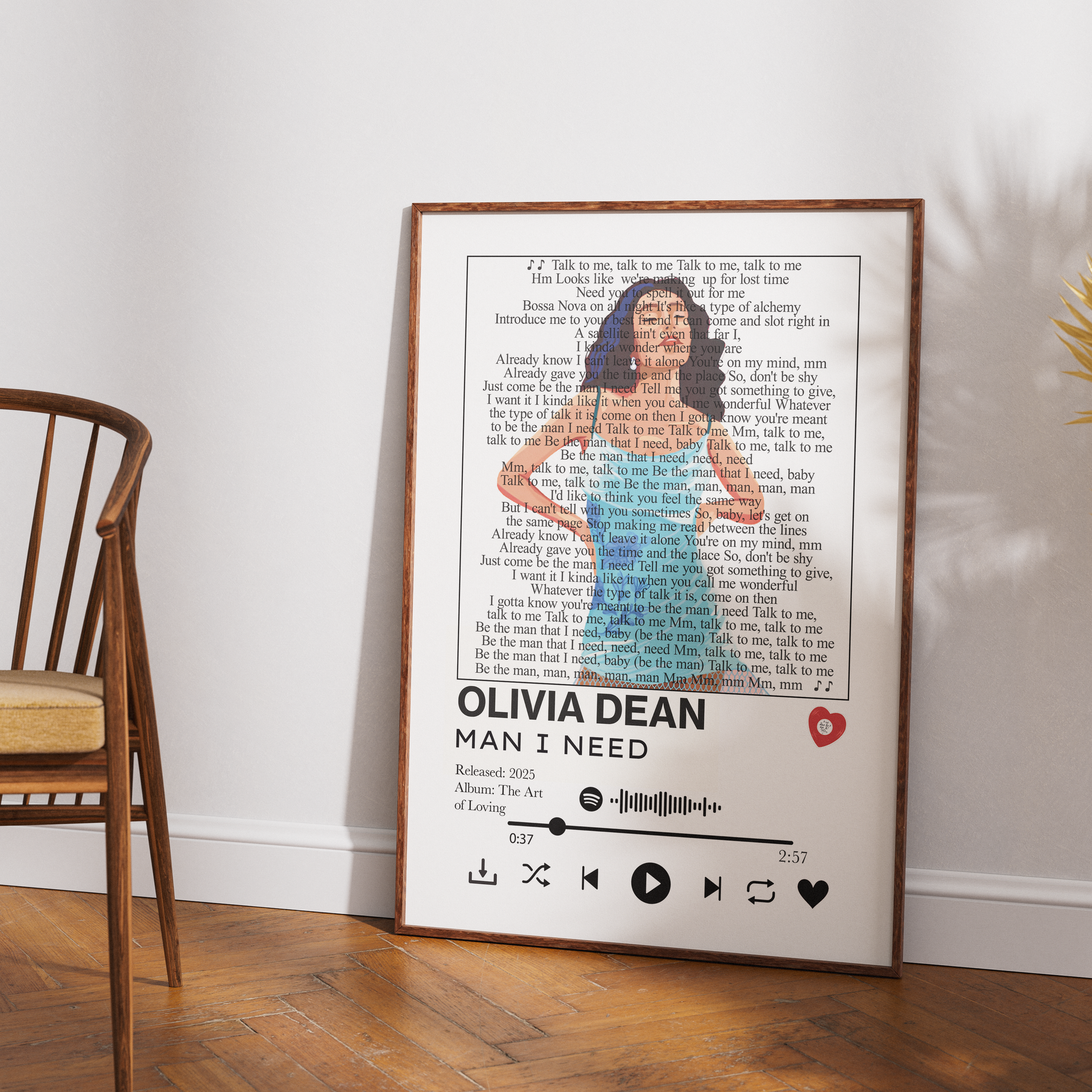 A framed personalized music poster of Olivia Dean's 'Man I Need' leaning against a white wall on a wooden floor, next to a wooden chair.