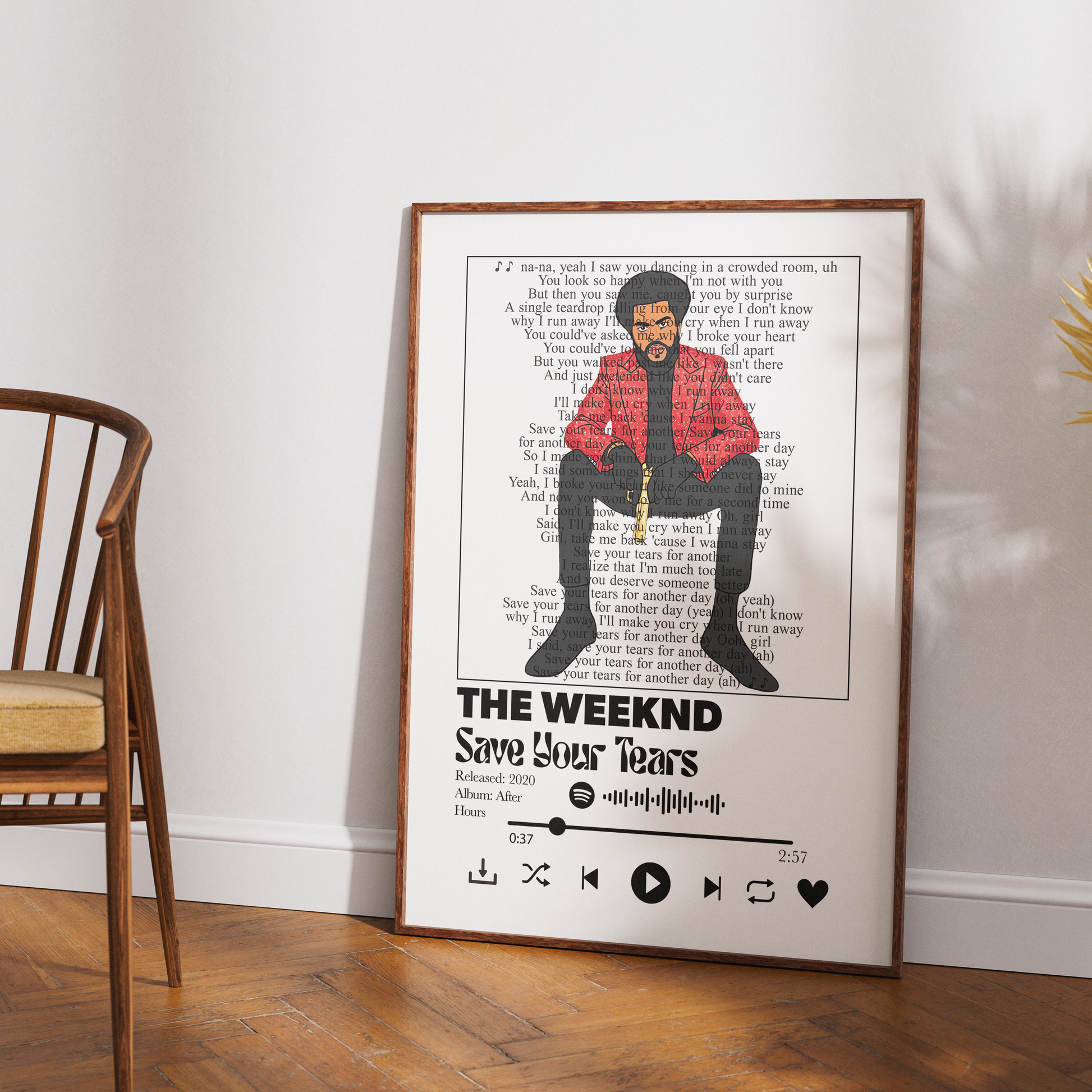 A framed music poster of The Weeknd's 'Save Your Tears' leaning against a white wall on a wooden floor, next to a wooden chair.