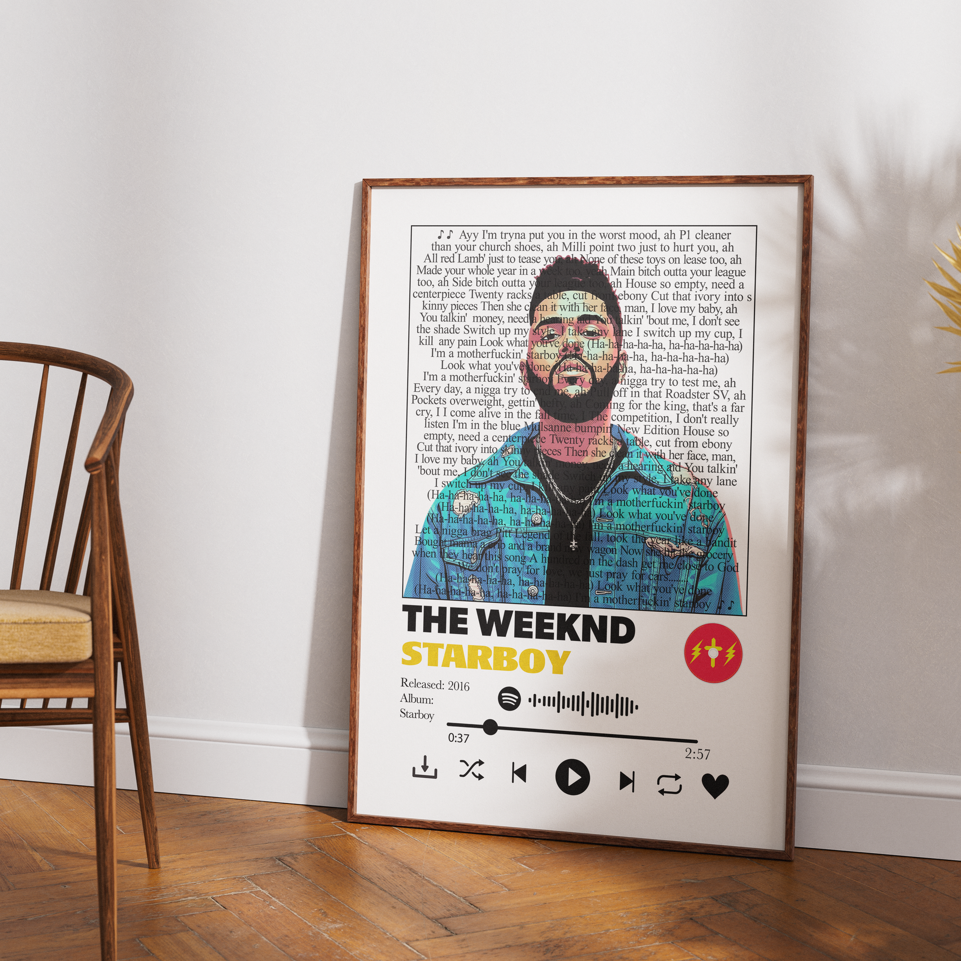 A framed music poster of The Weeknd's 'Starboy' leaning against a white wall on a wooden floor, next to a wooden chair.