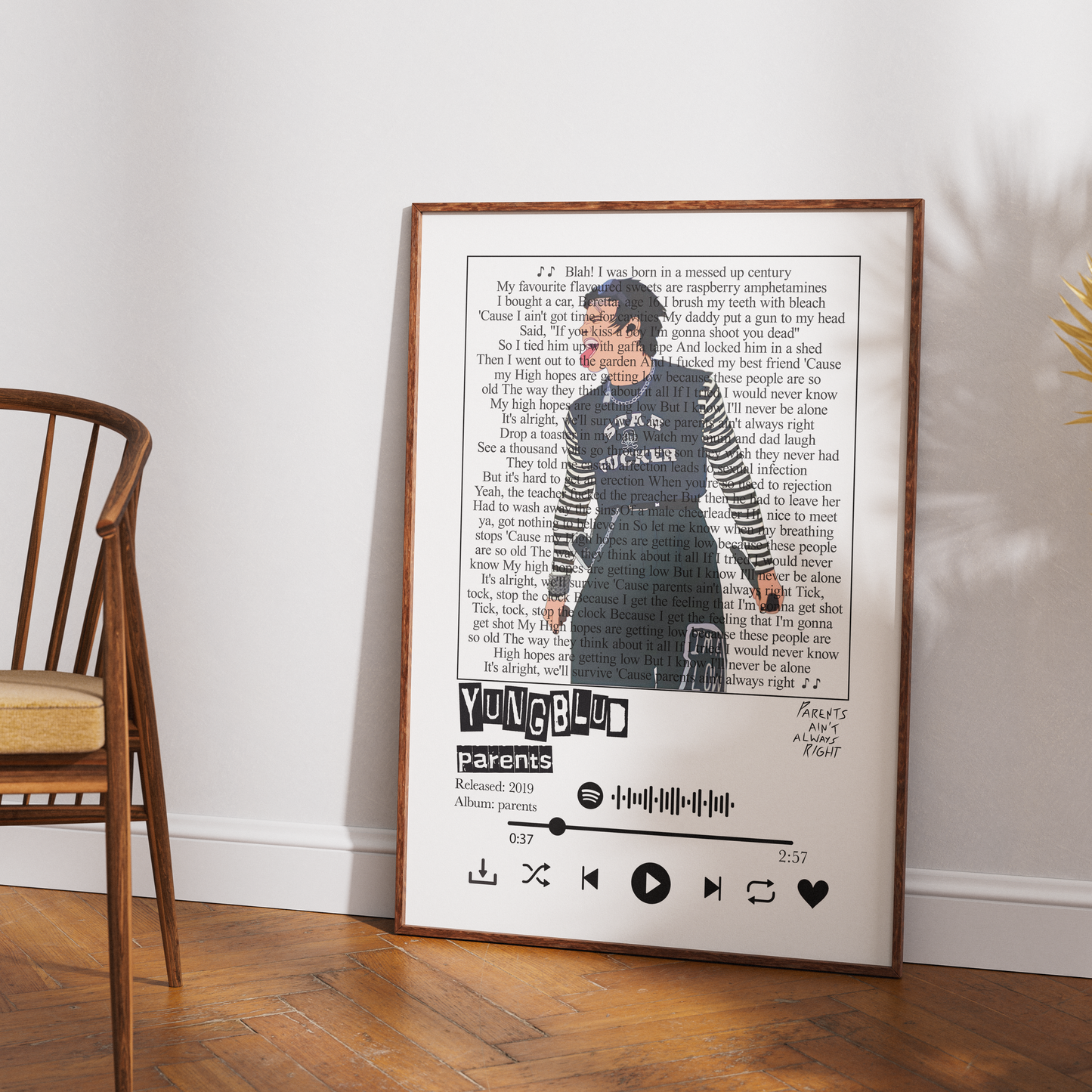 A framed custom music-themed art print leaning against a white wall on a wooden floor, next to a wooden chair. The print features a photo of a woman, song lyrics, the name 'YUNG61', and a custom playlist styled like a Spotify interface.